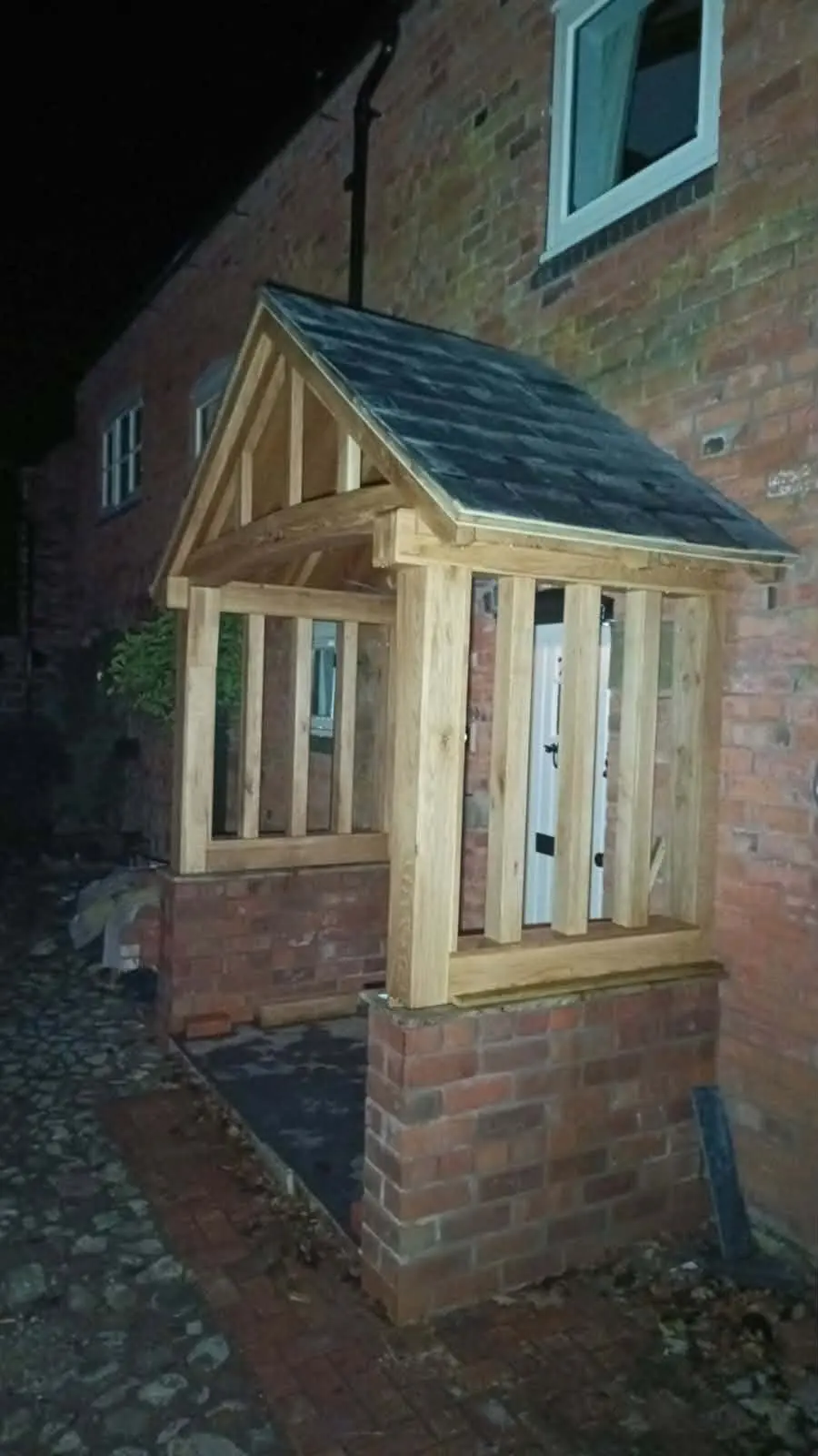 Timber porch frame with a pitched roof built against a brick wall, photographed after dark