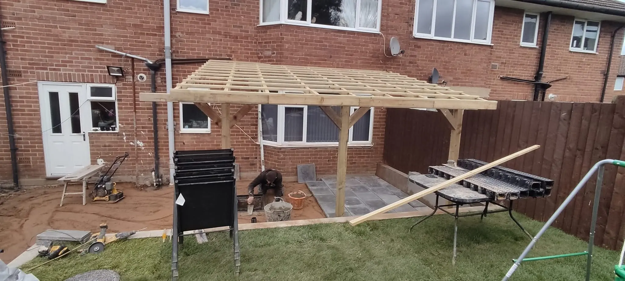 Timber roof framing attached to the back of a brick house, above a lawn and materials
